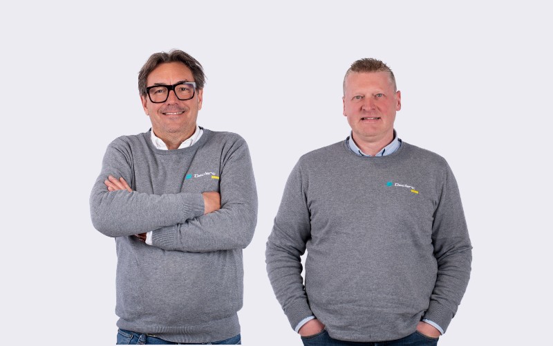 L'image montre un homme souriant, posant dans ce qui semble &ecirc;tre un magasin de v&eacute;los ou un atelier de r&eacute;paration. Il porte une chemise polo noire avec le logo "Declerck" brod&eacute; sur la poitrine. Il est appuy&eacute; contre un comptoir ou une table, avec une roue de v&eacute;lo visible &agrave; c&ocirc;t&eacute; de lui. Derri&egrave;re lui, il y a des plantes vertes et un mur bleu, ce qui donne une ambiance accueillante et professionnelle &agrave; l'endroit. L'homme semble &ecirc;tre un employ&eacute; ou le propri&eacute;taire du magasin.